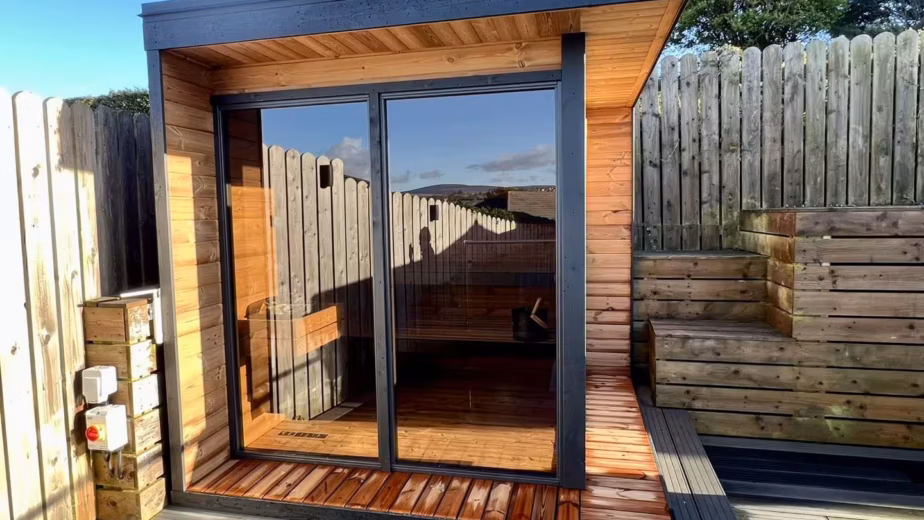 A modern wood-paneled Cube Sauna with a large panoramic glass window, recently installed in a residential garden in Onchan, Isle of Man, by Roberts Leisure.