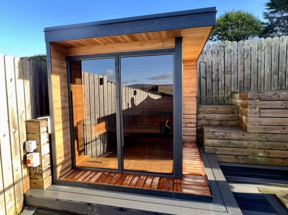 A modern wood-paneled Cube Sauna with a large panoramic glass window, recently installed in a residential garden in Onchan, Isle of Man, by Roberts Leisure.