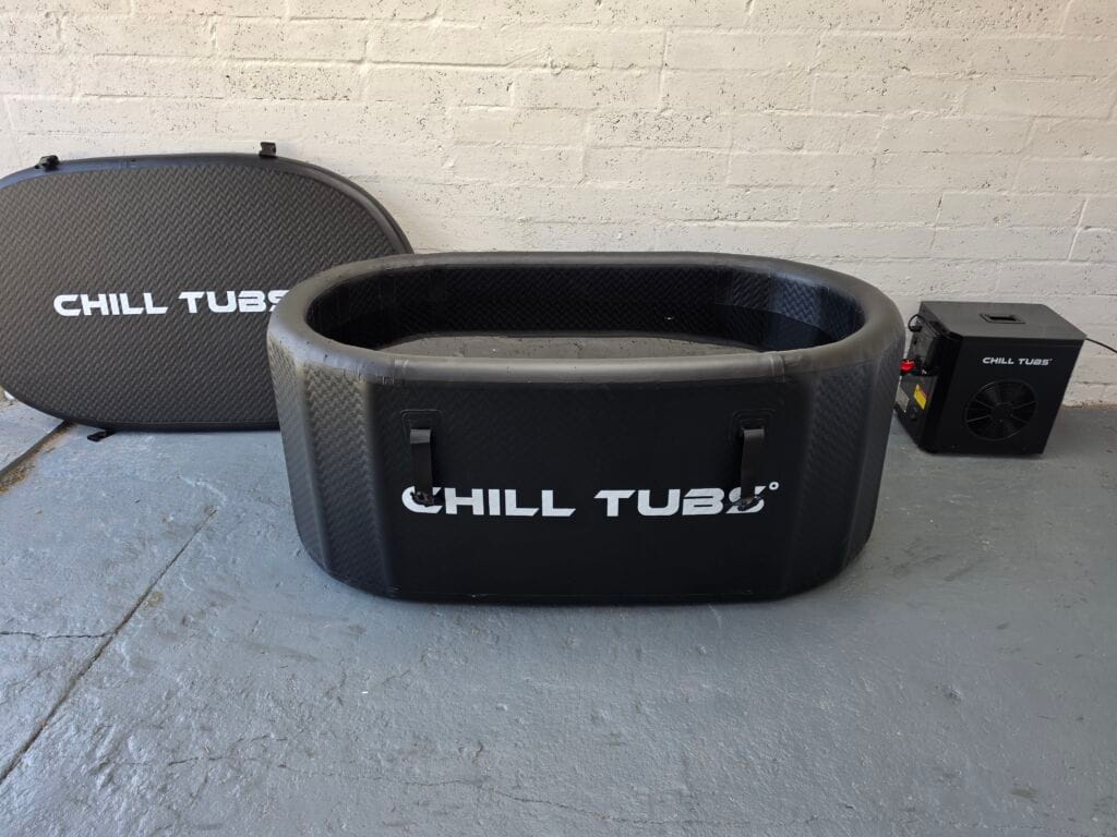 An open black Chill Tubs cold plunge bath, ready for use, with the black insulated lid leaning against a white brick wall behind it. An integrated electric chiller is connected to the right.