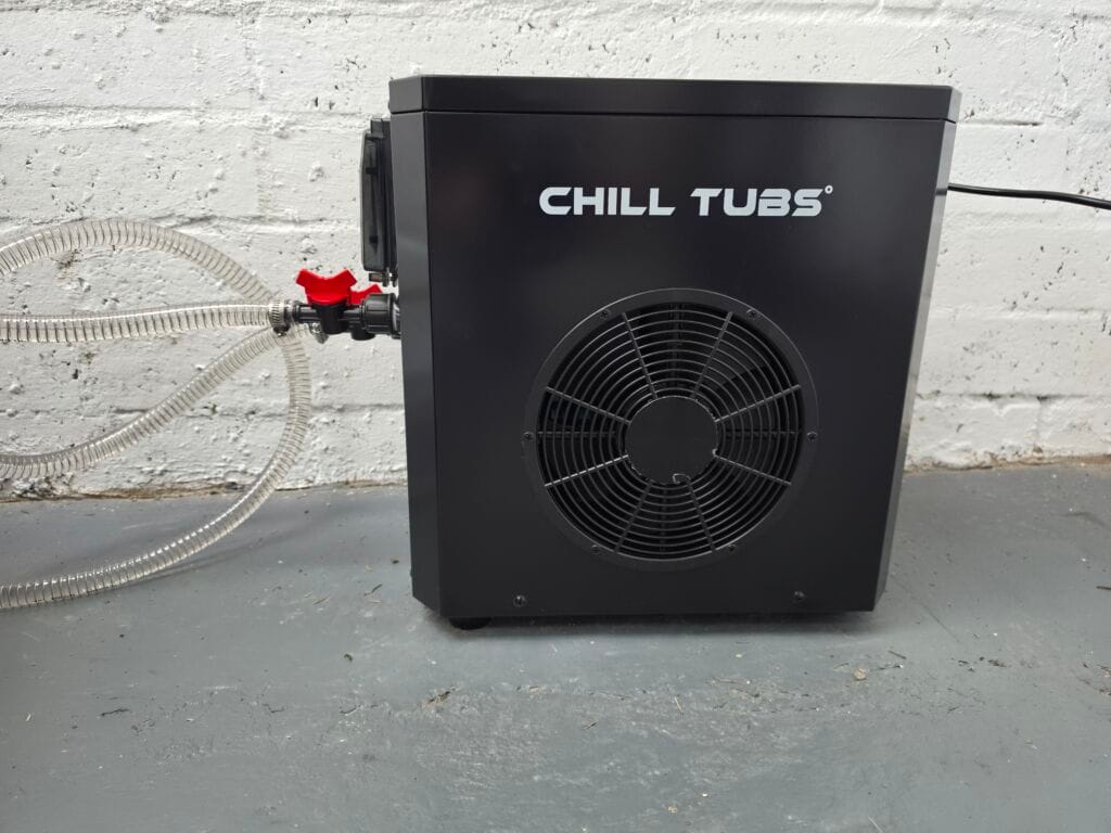 Close-up of a black, square Chill Tubs electric chiller unit with a circular fan grate. Two clear water hoses are connected to the left side with a red valve. It sits on a concrete floor against a white brick wall.