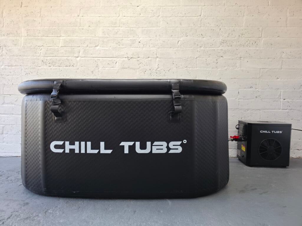 A black oval Chill Tubs cold plunge bath with its insulated cover buckled shut. To the right, a black electric chiller unit is connected via clear hoses. The setup sits on a gray concrete floor against a white painted brick wall.