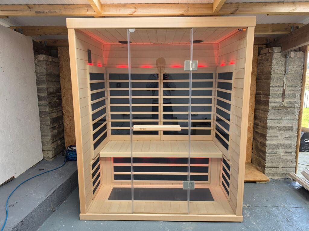 Interior view of a light-wood Tylö T825H infrared sauna cabin with red LED mood lighting and dark heating panels, placed within an under-construction wellness space on a concrete floor. A plywood wall and concrete block column are visible. A blue air hose rests on the ground. The glass door reflects the photographer.
