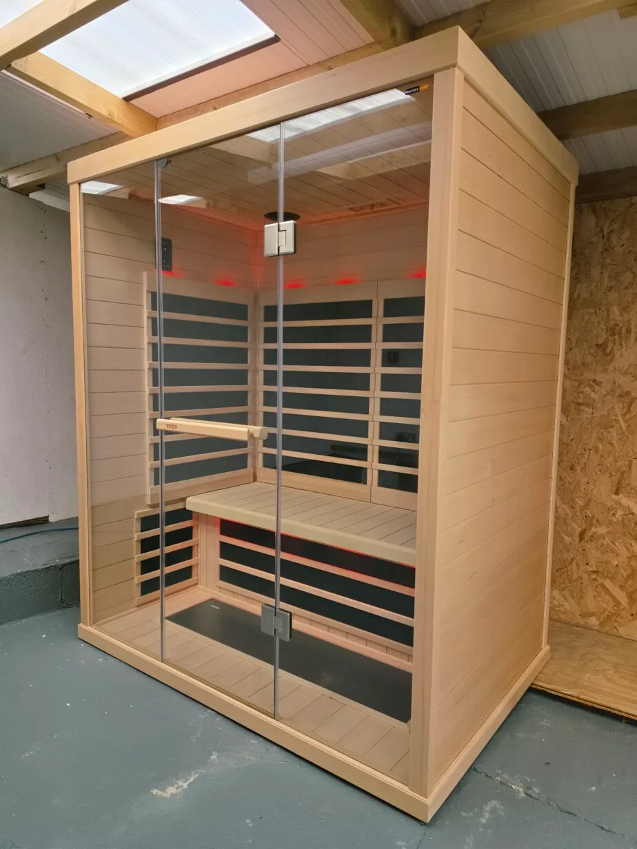 External front view of a large Tylö T825H infrared sauna cabin with a full glass front and wooden frame, placed within a garage featuring concrete block pillars and an OSB wooden wall. The photographer's reflection is clearly visible in the glass. Red LED mood lighting glows within the sauna. A blue air hose is on the concrete floor.