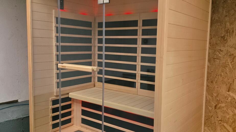 External front view of a large Tylö T825H infrared sauna cabin with a full glass front and wooden frame, placed within a garage featuring concrete block pillars and an OSB wooden wall. The photographer's reflection is clearly visible in the glass. Red LED mood lighting glows within the sauna. A blue air hose is on the concrete floor.