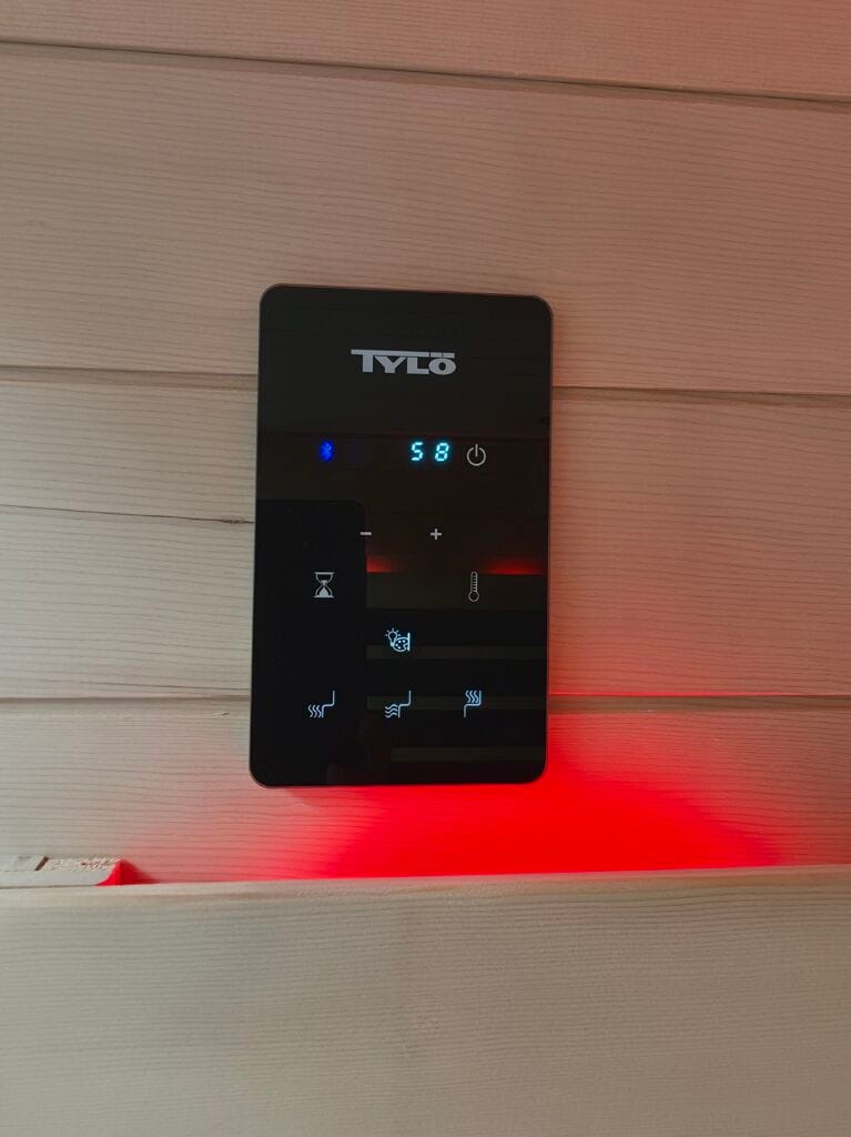 Close-up of a black glass Tylö touchscreen control panel mounted on light wooden sauna wall panels. The digital screen displays a blue Bluetooth icon and a temperature reading of 58. Icons for a timer, lighting, and heat settings are visible. A bright red LED mood light glows directly beneath the panel.