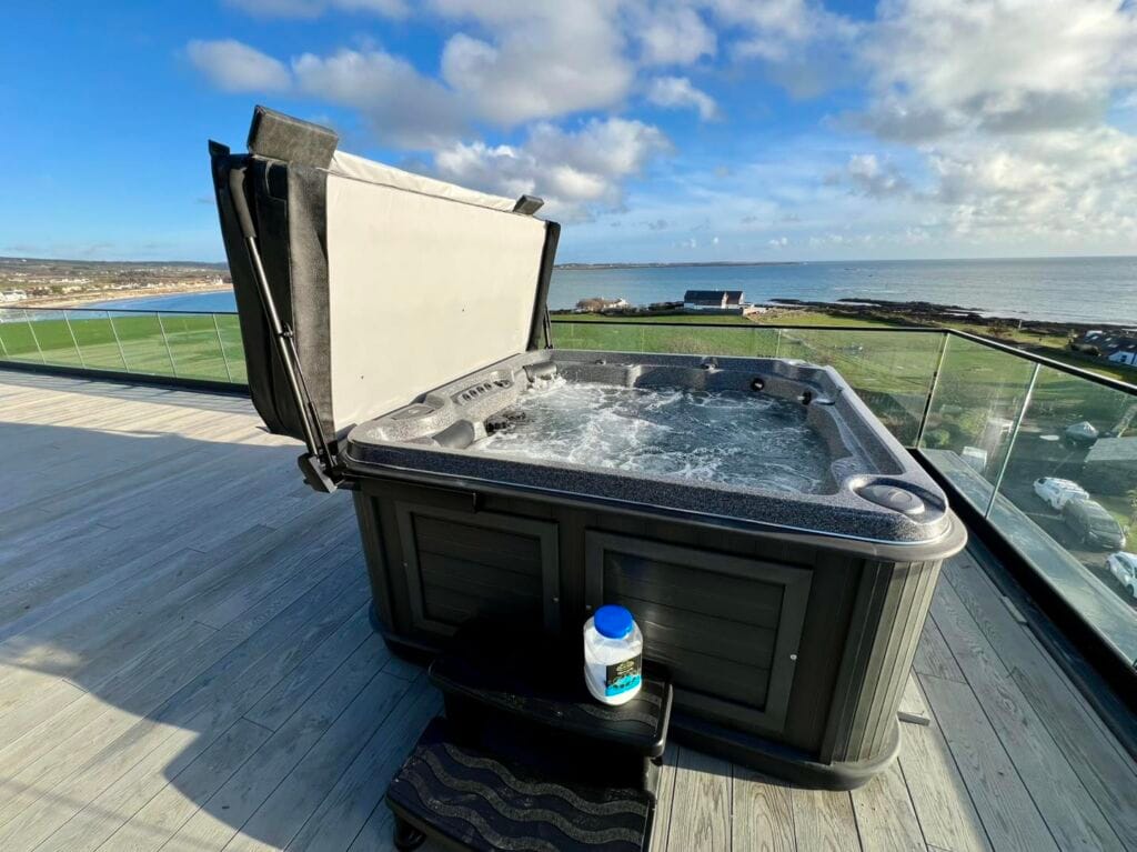 Side view of a dark grey Arctic Spas Mustang hot tub with the cover lifted open via a mechanical arm, sitting on a spacious modern balcony overlooking the coast in Port St Mary, Isle of Man, Roberts Leisure