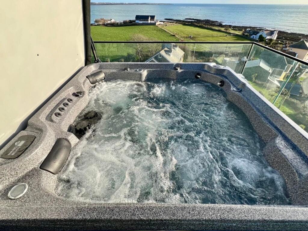 A bubbling grey hot tub installed on a modern glass-fronted balcony, overlooking a stunning coastal landscape of green fields and the sea on the IOM, Roberts Leisure