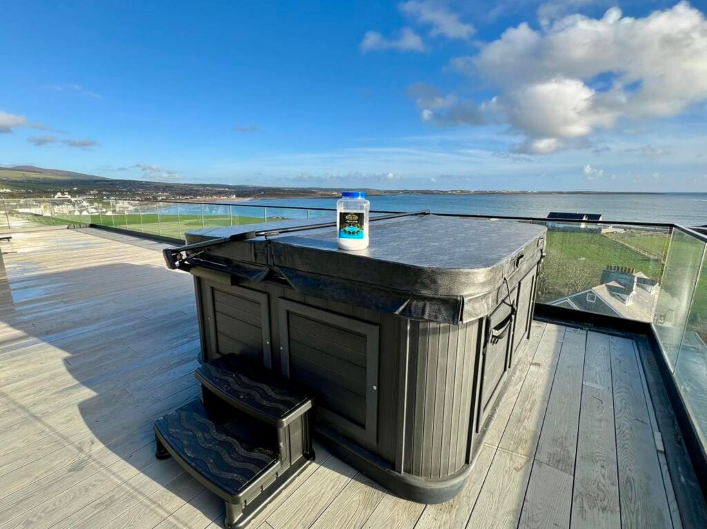 A dark grey hot tub securely closed with a heavy-duty insulated thermal cover, sitting on a modern balcony overlooking the coast in Port St Mary, IOM. A container of spa water care salt rests on top, Roberts Leisure