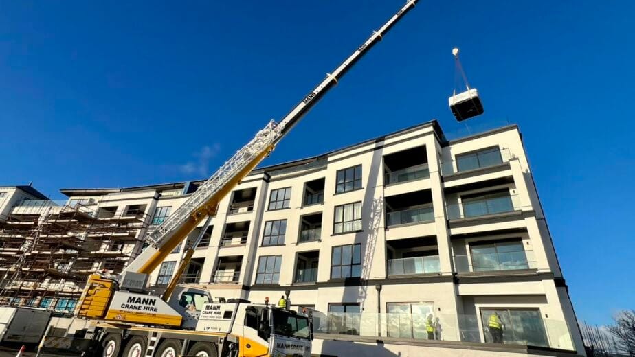 600295786 1789502552228213 707702851253519A large yellow and white Mann Crane Hire mobile crane lifting a wrapped hot tub high in the air, positioned to land on the top balcony of a modern multi-story apartment building under construction in the IOM, Roberts Leisure.
