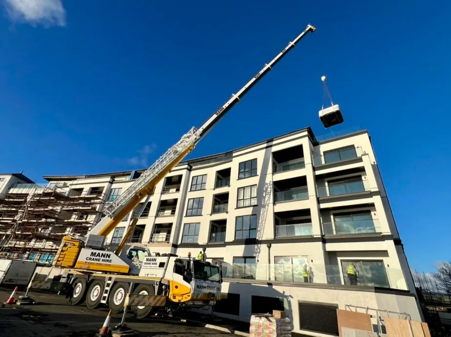 600295786 1789502552228213 707702851253519A large yellow and white Mann Crane Hire mobile crane lifting a wrapped hot tub high in the air, positioned to land on the top balcony of a modern multi-story apartment building under construction in the IOM, Roberts Leisure.