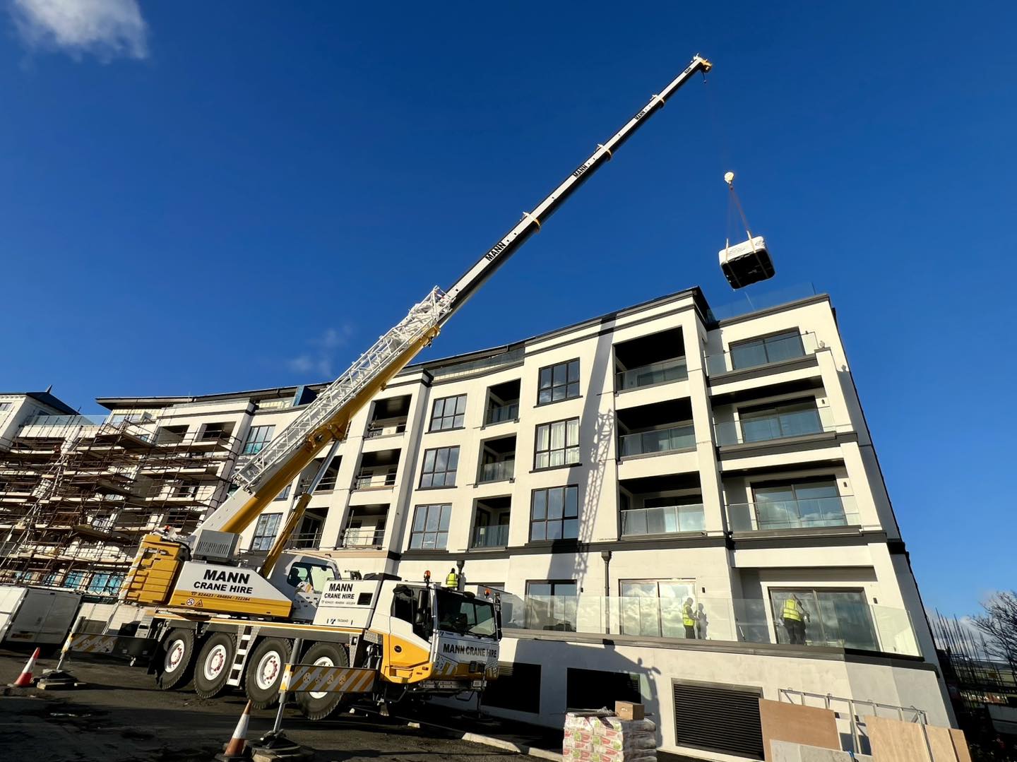 600295786 1789502552228213 707702851253519A large yellow and white Mann Crane Hire mobile crane lifting a wrapped hot tub high in the air, positioned to land on the top balcony of a modern multi-story apartment building under construction in the IOM, Roberts Leisure.