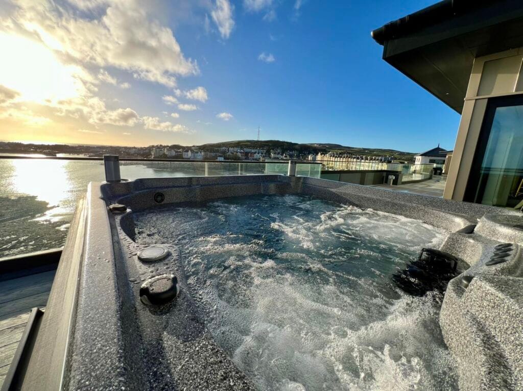 Wide angle view of a bubbling grey hot tub on a penthouse balcony with glass railings, looking out over the sunlit town, coastline, and harbor of Port St Mary on the Isle of Man, Roberts Leisure