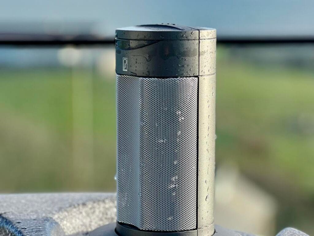 Close-up of a cylindrical pop-up Bluetooth waterproof hot tub speaker covered in water droplets, integrated into the shell of a premium spa against a blurred coastal background in the IOM, Roberts Leisure