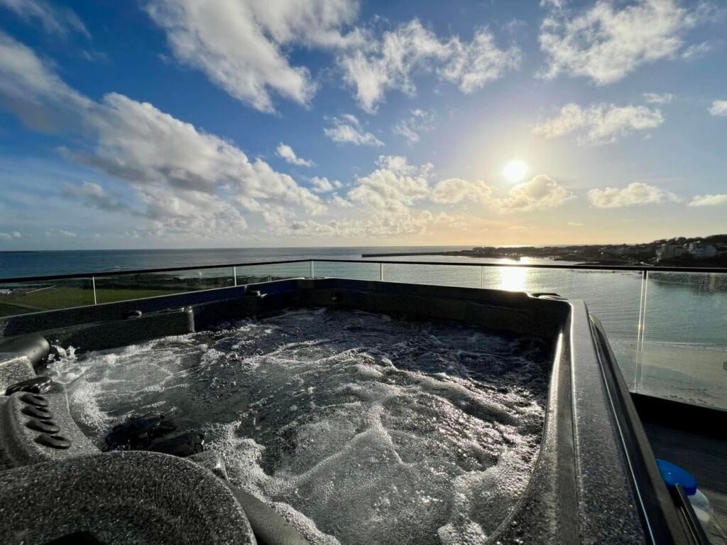 A bubbling dark grey hot tub on a glass-fronted balcony, beautifully illuminated by the late afternoon sun setting over the ocean and harbour in Port St Mary, IOM, Roberts Leisure