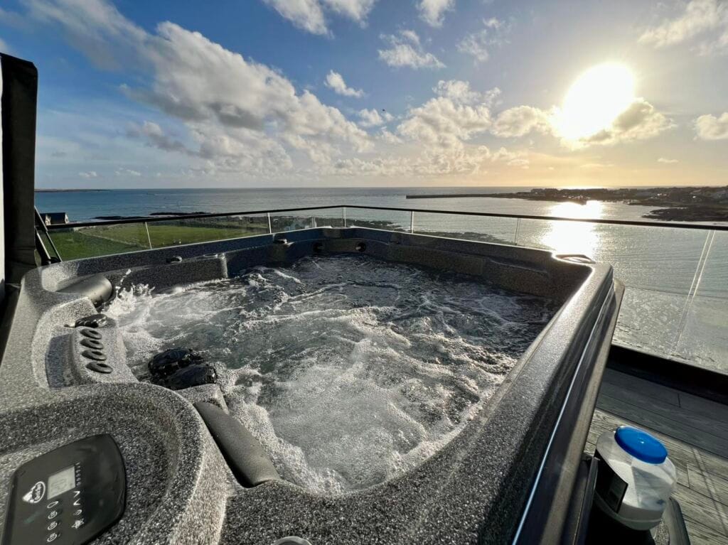 A bubbling grey hot tub operating on a glass-fronted balcony, featuring a spectacular sunlit view over the ocean and harbor in Port St Mary, Isle of Man, Roberts Leisure.