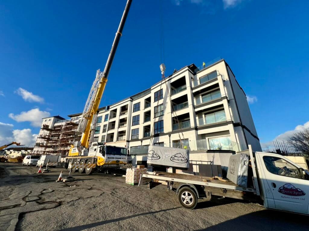 A large mobile crane lifting a wrapped hot tub from a Roberts Leisure flatbed truck onto the balcony of a modern multi-story apartment building under a clear blue sky on the Isle of Man