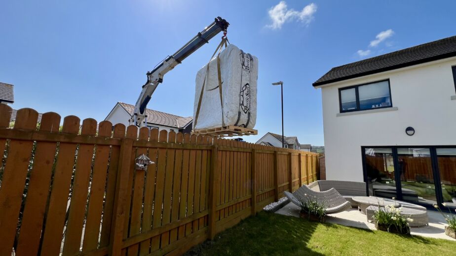 A crane lifting a securely packaged Arctic Spas hot tub high over a wooden garden fence into a residential property on the Isle of Man under a bright, sunny sky.