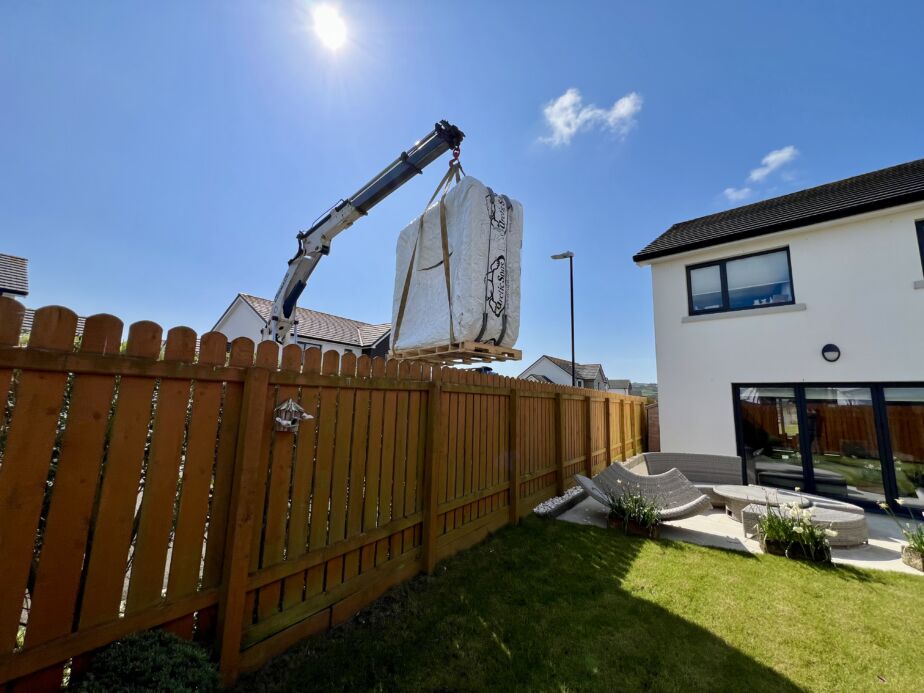 A crane lifting a securely packaged Arctic Spas hot tub high over a wooden garden fence into a residential property on the Isle of Man under a bright, sunny sky.