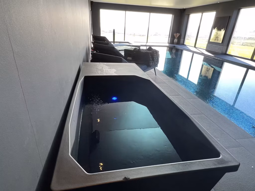 An angled wide shot of the Chill Tub Lite ice bath on a grey slate deck, overlooking luxury lounge chairs and a glassy indoor swimming pool with floor-to-ceiling windows showing the Manx countryside.