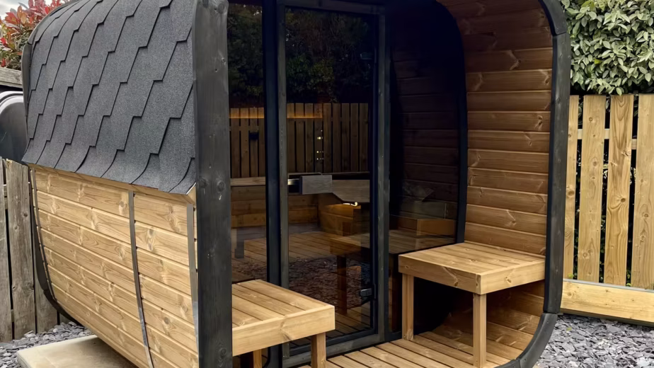 A premium freestanding Yakia 200L outdoor cube-barrel sauna made of natural Thermowood with black shingle roofing, integrated front canopy porch, and twin wooden cooling benches on a slate gravel patio.