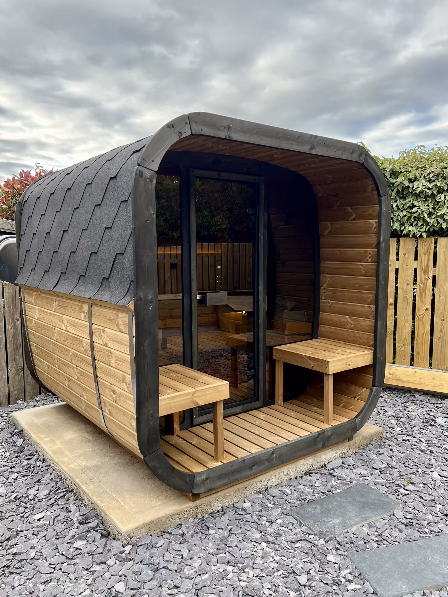 A premium freestanding Yakia 200L outdoor cube-barrel sauna made of natural Thermowood with black shingle roofing, integrated front canopy porch, and twin wooden cooling benches on a slate gravel patio.
