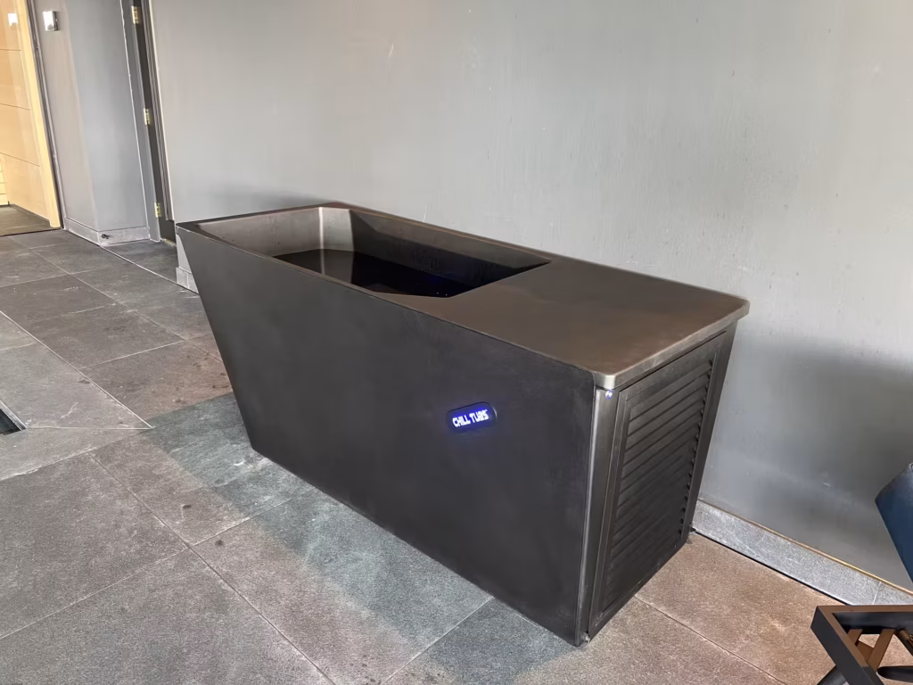 A full view of the freestanding black Chill Tub Lite cold plunge unit on dark grey flagstones, showing its minimalist rectangular profile, integrated maintenance grille, and illuminated blue logo badge.