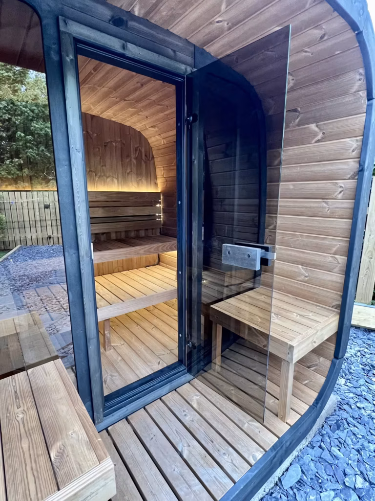 A close-up angled view of the open tinted glass door on a Yakia 200L outdoor sauna, revealing a warmly lit interior with split-level wooden benches and subtle LED backlighting.