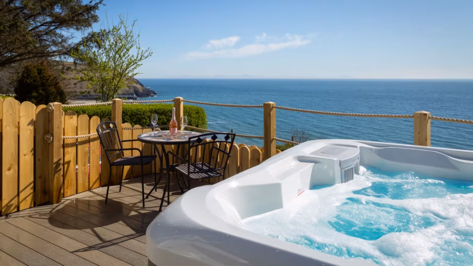 A bubbling AquaSolus Vacation Cypress hot tub installed on a wooden deck with a patio table and wine glasses, overlooking a stunning bright blue ocean view in Laxey, Isle of Man.