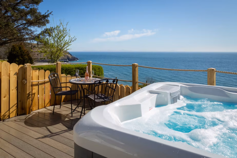 A bubbling AquaSolus Vacation Cypress hot tub installed on a wooden deck with a patio table and wine glasses, overlooking a stunning bright blue ocean view in Laxey, Isle of Man.