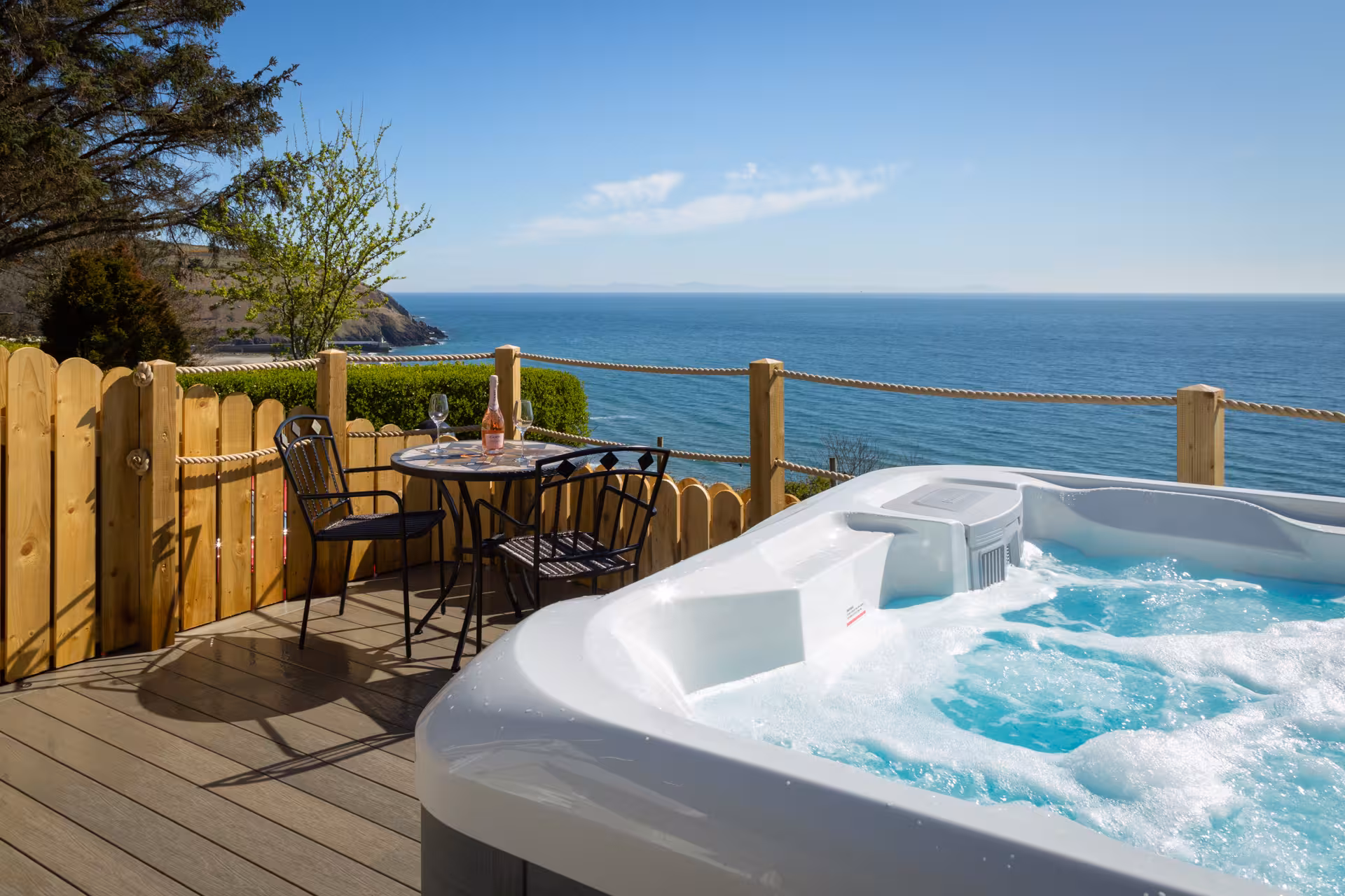 A bubbling AquaSolus Vacation Cypress hot tub installed on a wooden deck with a patio table and wine glasses, overlooking a stunning bright blue ocean view in Laxey, Isle of Man.
