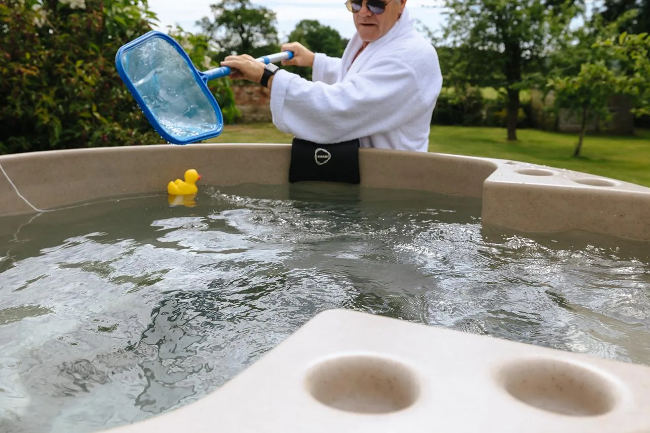 A close-up looking into a beige Kirami wood-fired hot tub with a floating yellow rubber duck, as a man in a white bathrobe uses a blue pool skimmer net to clear debris from the water in an Isle of Man garden.