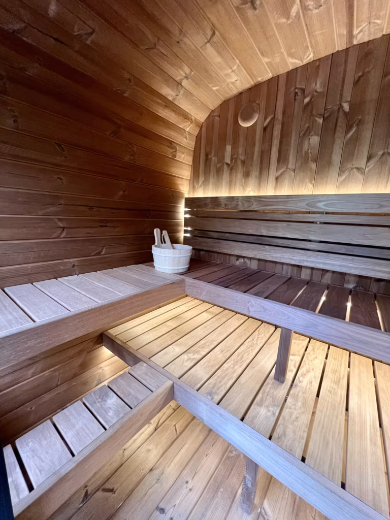 The interior of a Yakia 200L outdoor sauna featuring multi-level L-shaped Thermo-Aspen benches, warm integrated LED backlighting, and a traditional wooden bucket and ladle.
