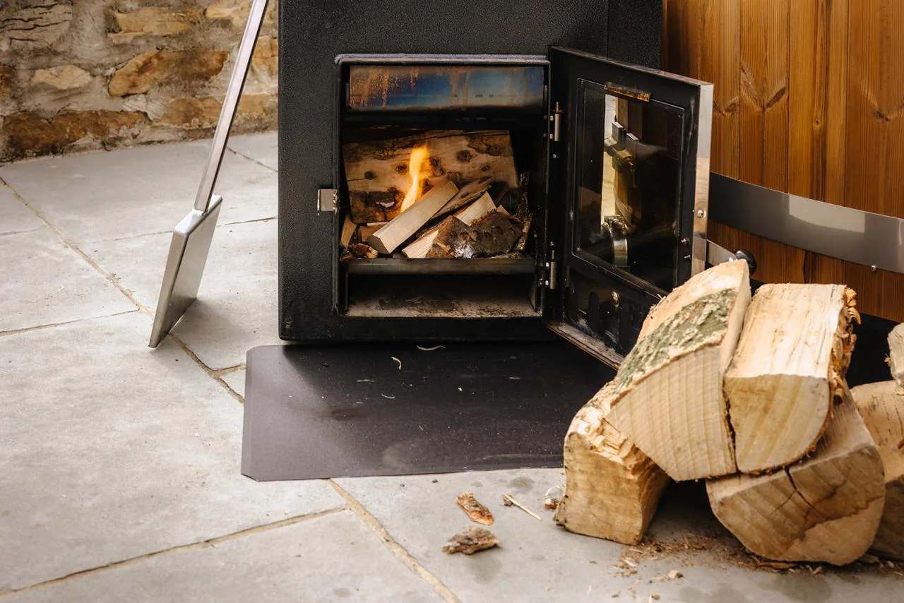A close-up of an external black Kirami hot tub heater stove with its glass door open, showing logs burning brightly inside on a stone patio with split firewood stacked beside it on the Isle of Man.