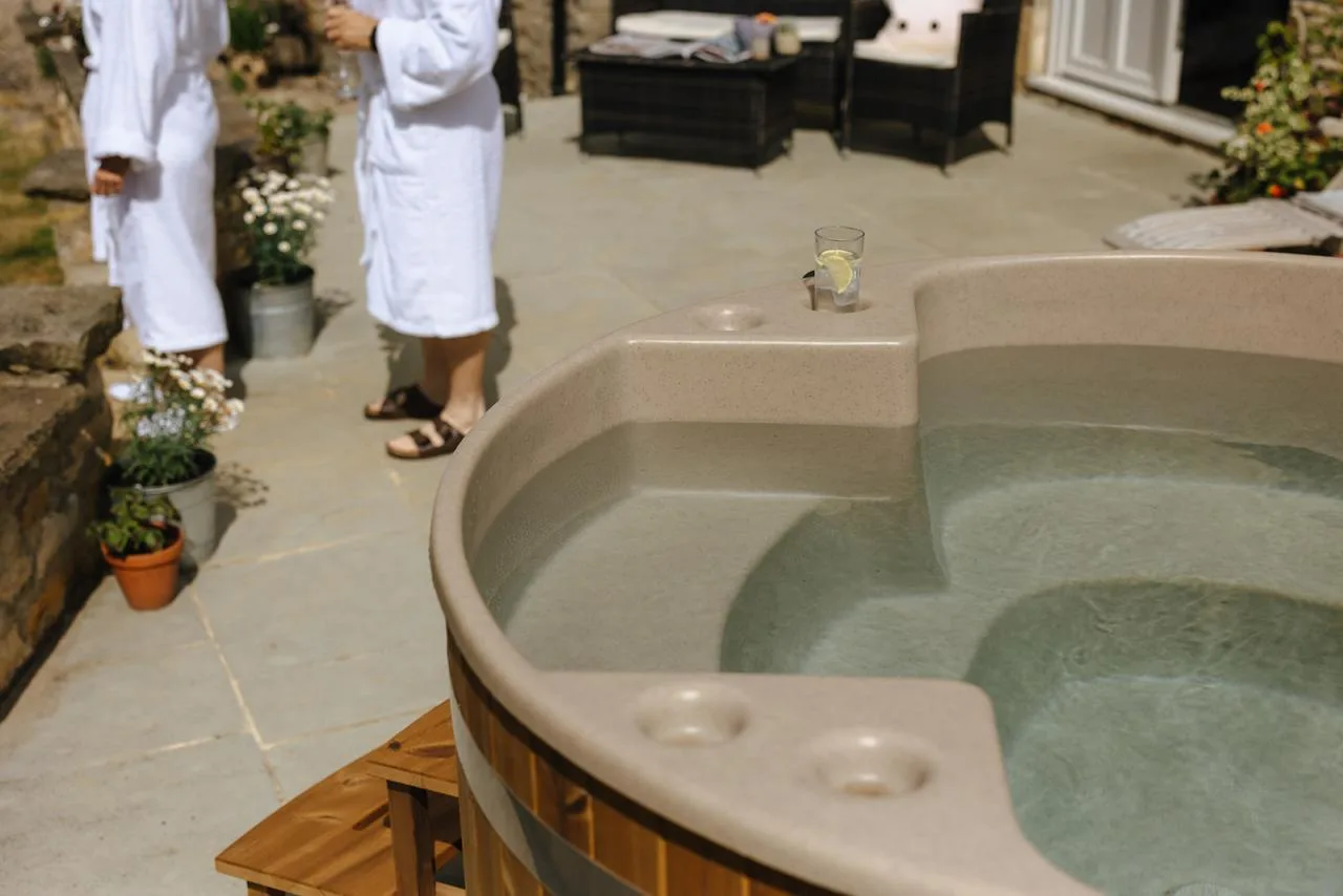 A close-up view of a beige molded Kirami wood-fired hot tub with a drink on the integrated cup holder console, positioned on a stone patio with people in white bathrobes standing in the background on the Isle of Man.