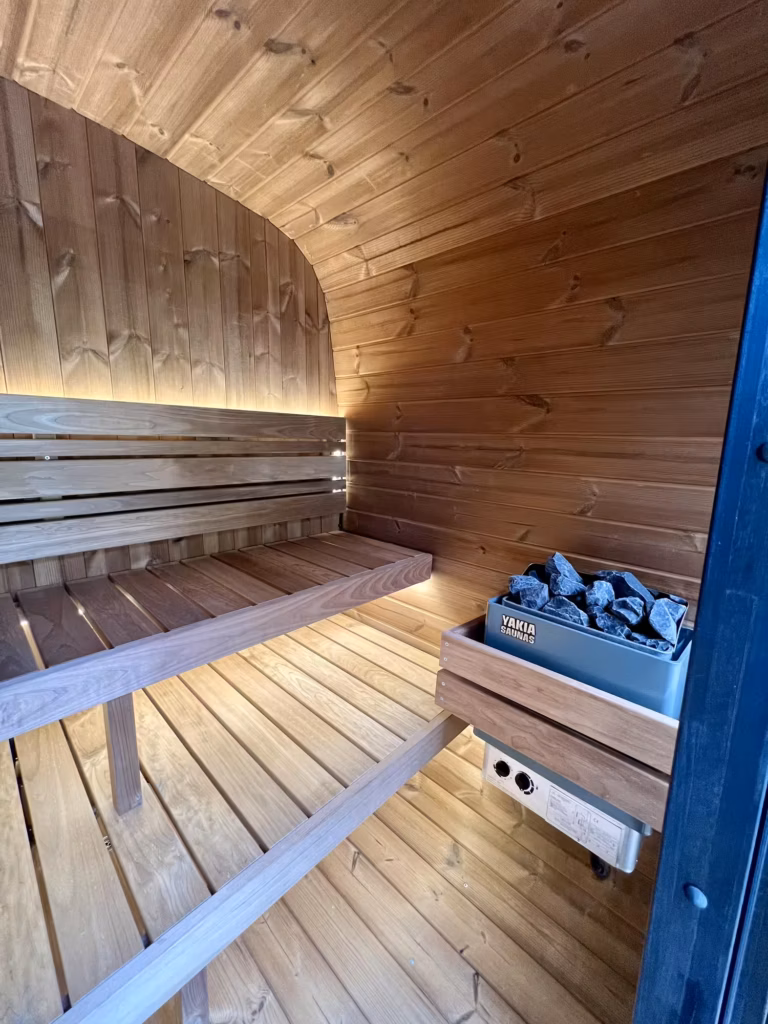 Interior view of a Yakia 200L outdoor sauna showing the wall-mounted Yakia Saunas electric heater filled with dark volcanic rocks, protected by a timber safety guard next to Thermo-Aspen benches.