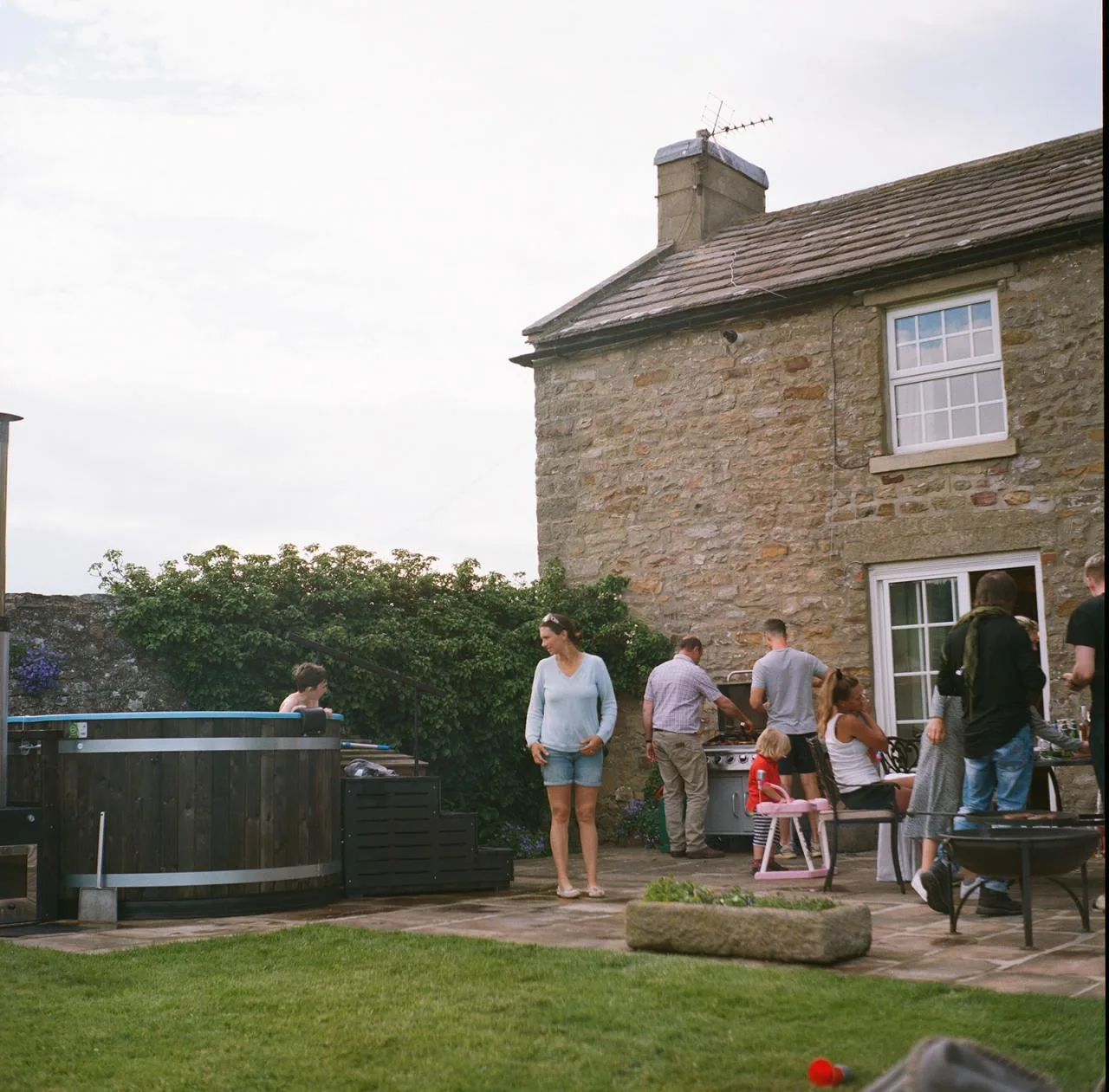A lively garden party on a stone patio outside a traditional stone house on the Isle of Man, with a dark wood Kirami wood-fired hot tub on the left and a group gathering around a barbecue on the right.