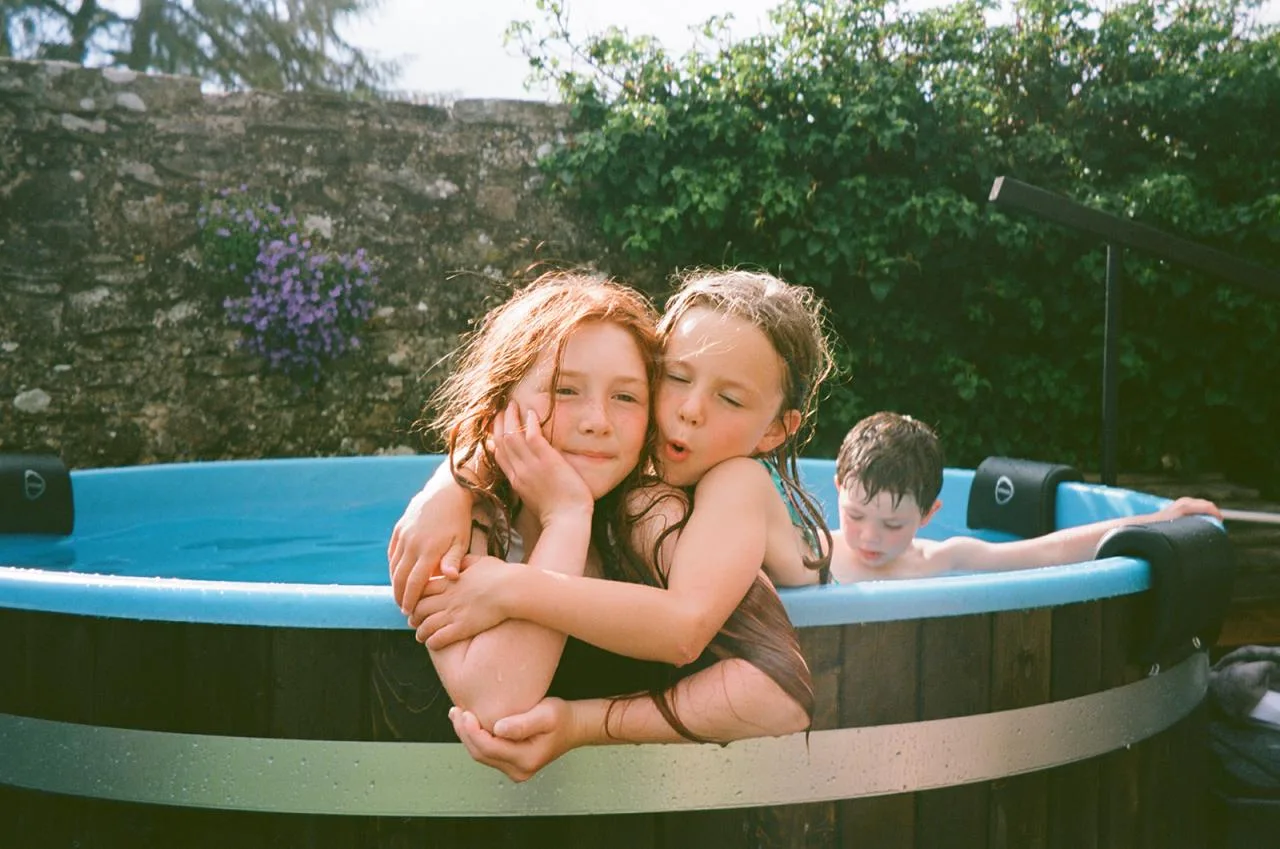 Two young girls smiling and hugging over the rim of a dark wood Kirami wood-fired hot tub with a light blue liner and black headrests, while a young boy plays in the background on the Isle of Man.