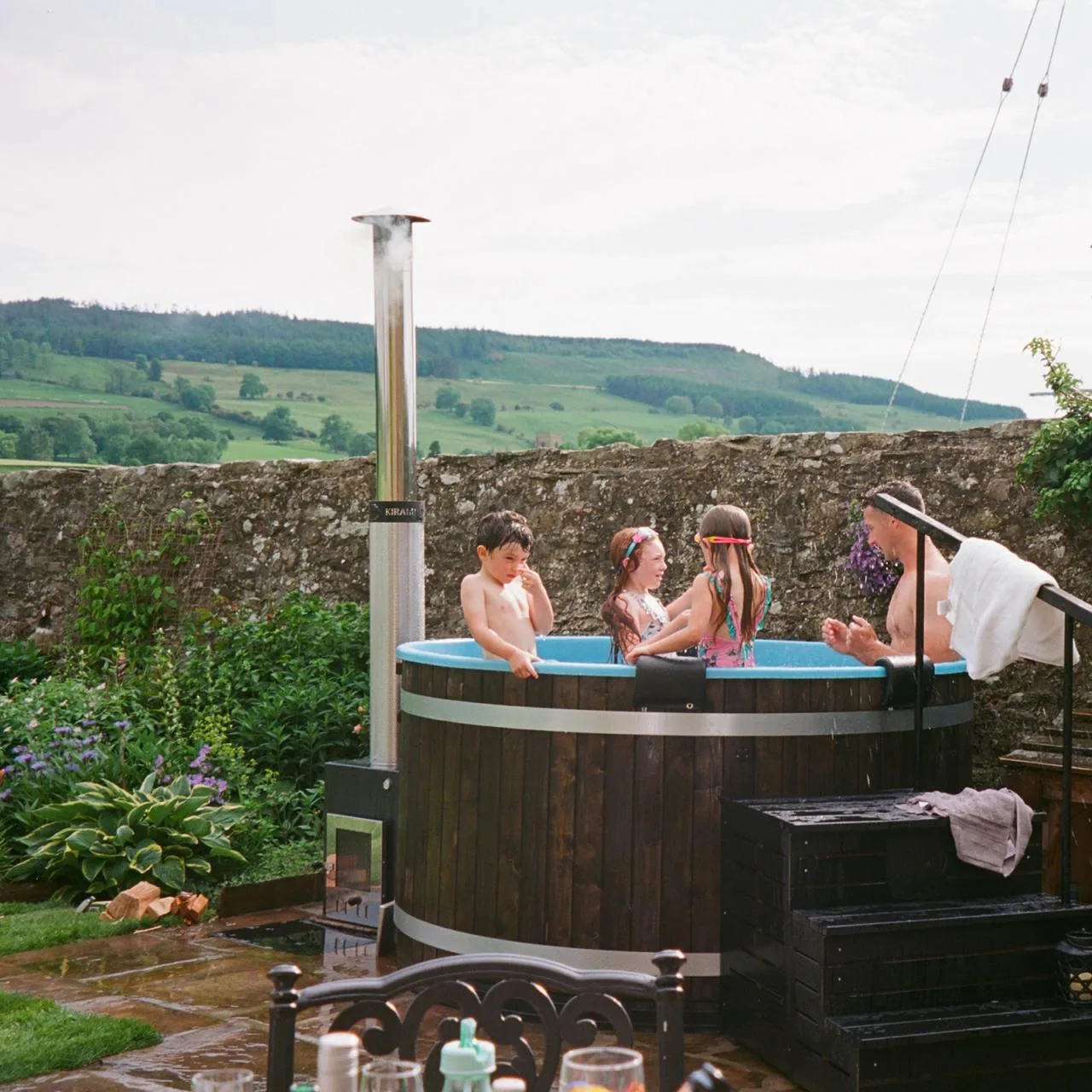 A father and three children enjoying a soak in a dark timber Kirami wood-fired hot tub with a light blue liner on a wet stone patio, looking out over a rustic stone wall toward green rolling hills on the Isle of Man.
