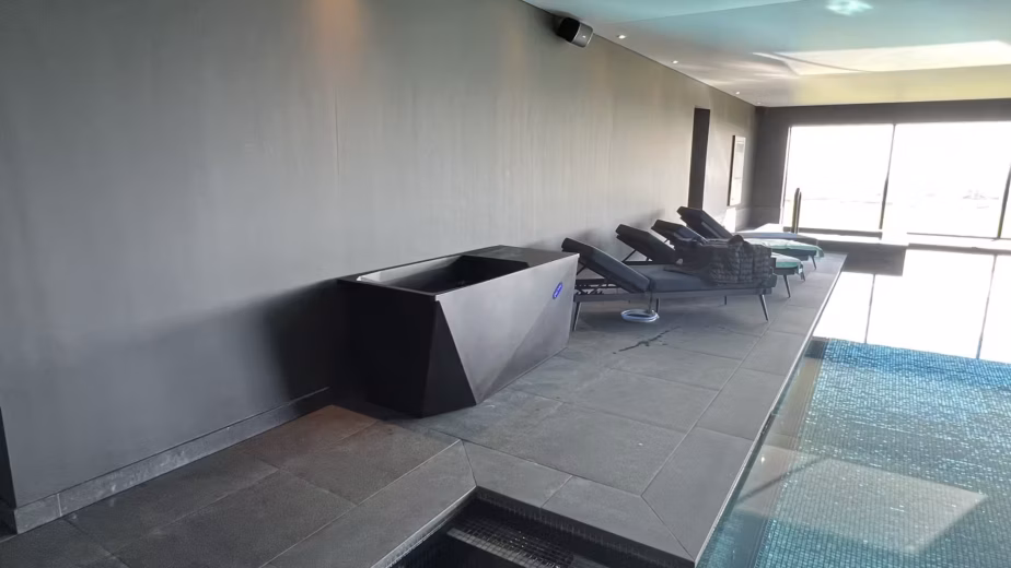 A sleek, geometric black Chill Tub Lite cold plunge pool situated on a dark tiled walkway next to a heated indoor swimming pool and lounge chairs.