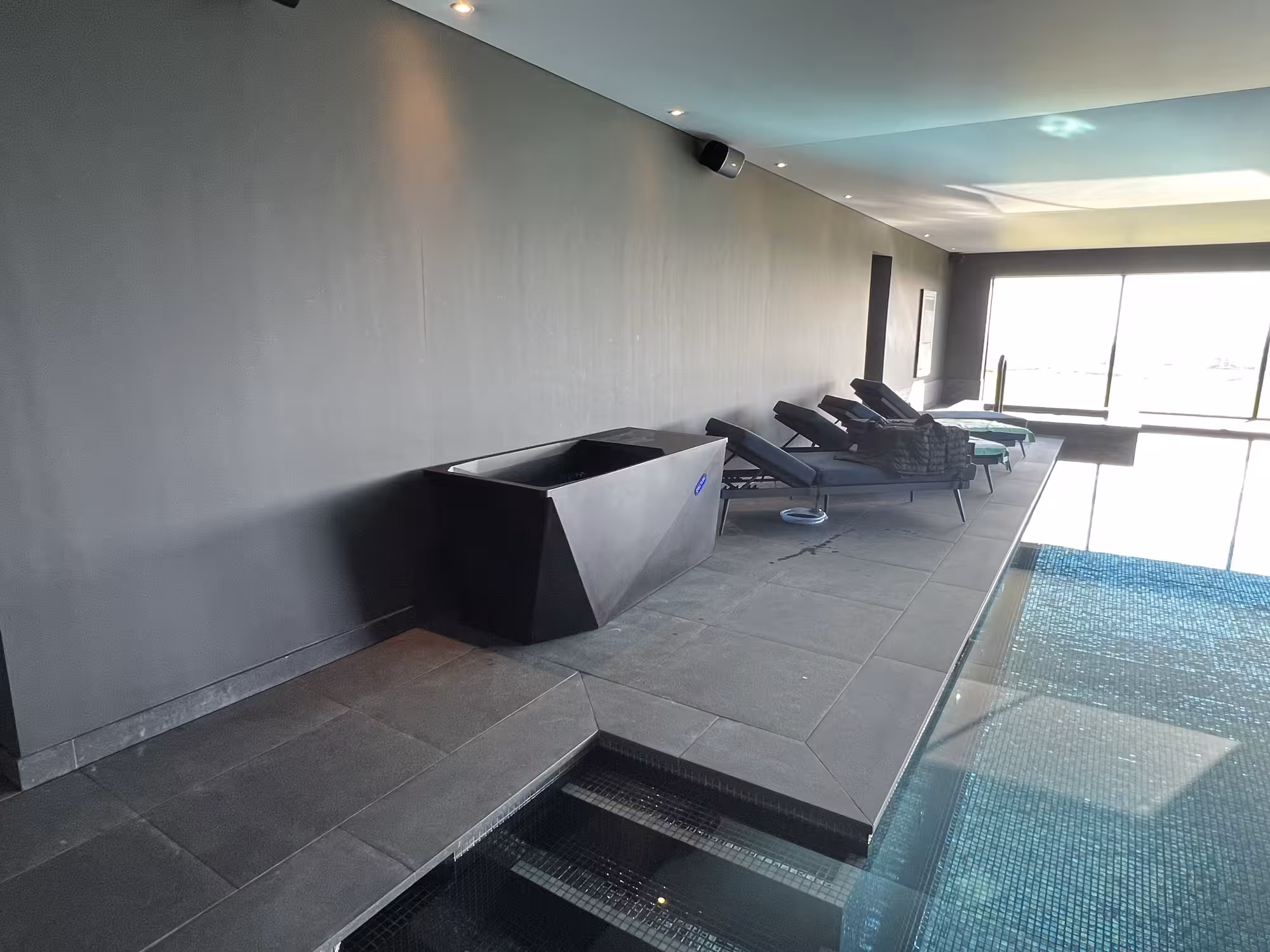 A sleek, geometric black Chill Tub Lite cold plunge pool situated on a dark tiled walkway next to a heated indoor swimming pool and lounge chairs.