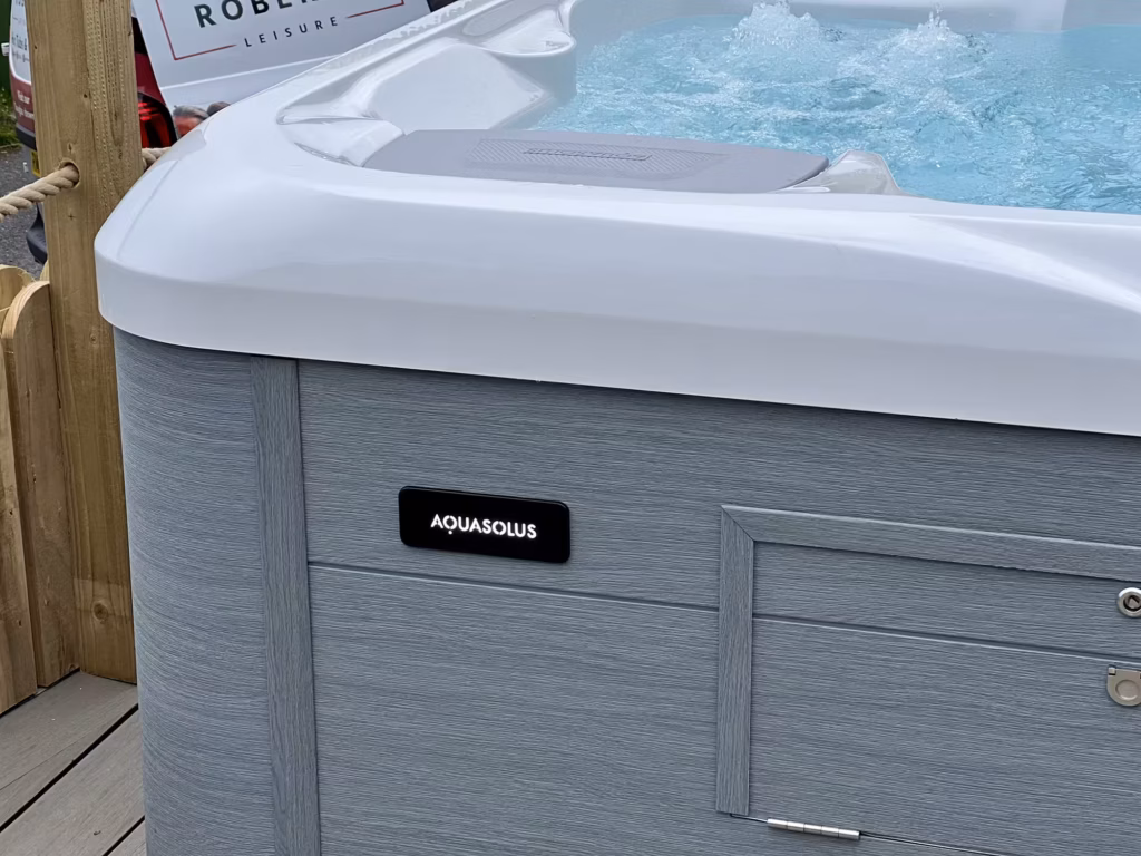A close-up of the black AquaSolus logo badge mounted on the textured grey exterior cabinet of a bubbling hot tub installed in the Isle of Man.