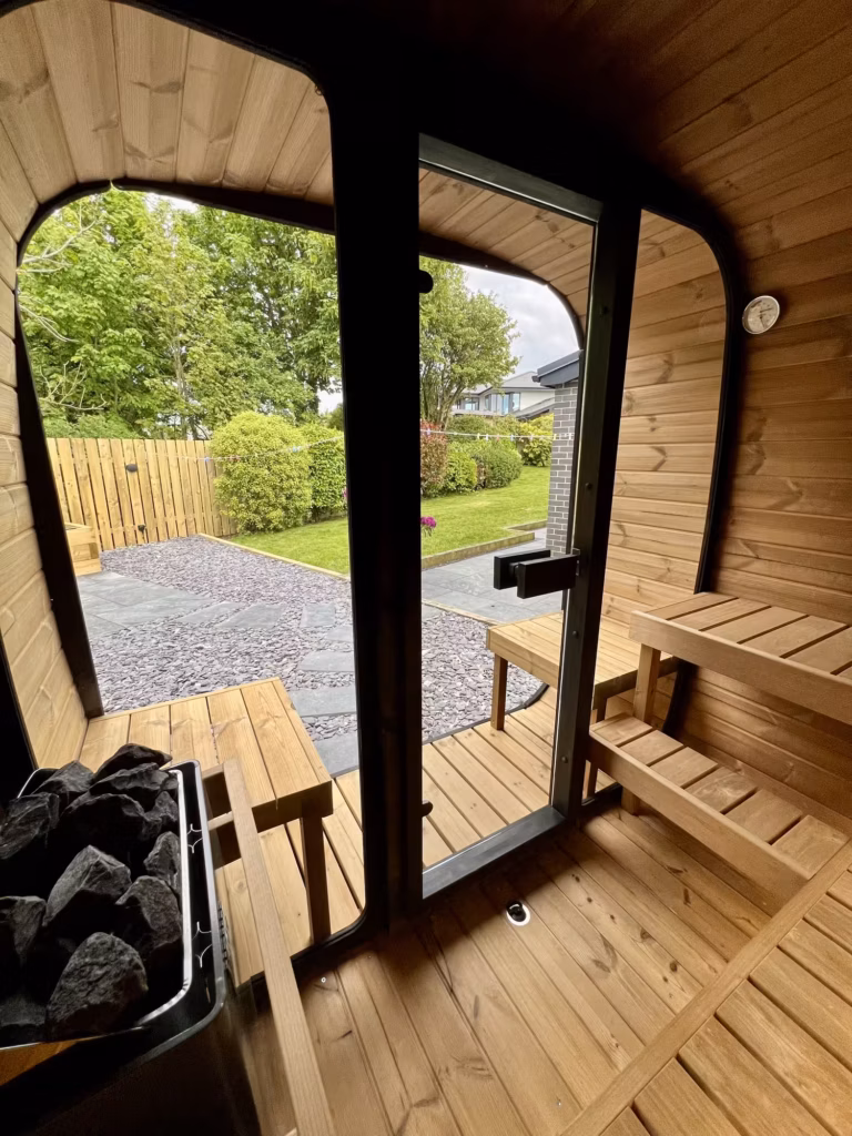 An interior perspective from inside a Yakia 200L outdoor sauna looking through the glass door onto a landscaped garden patio with grey slate stone and green lawn.