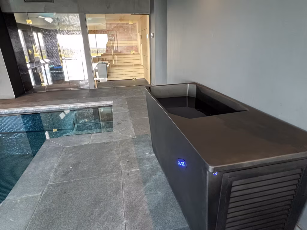 A black Chill Tub Lite cold plunge pool positioned on a stone tile floor alongside an indoor swimming pool, with a glass-fronted traditional sauna visible in the background.