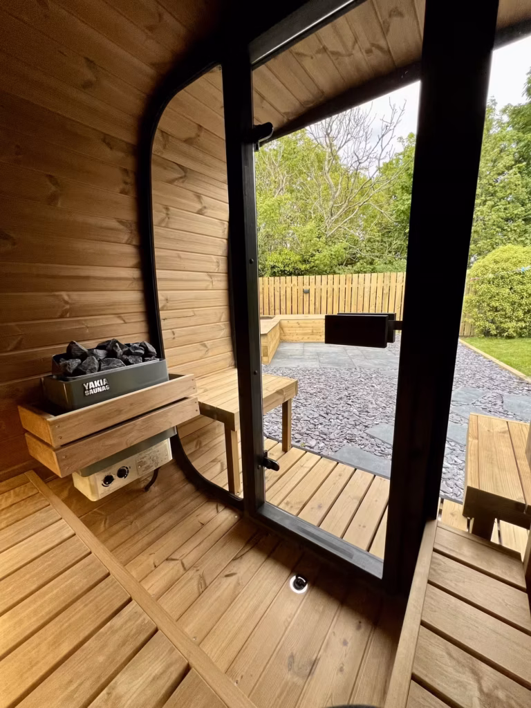 An interior view from a Yakia 200L outdoor sauna looking through the open glass door toward a private timber-fenced garden terrace with slate stone chips.
