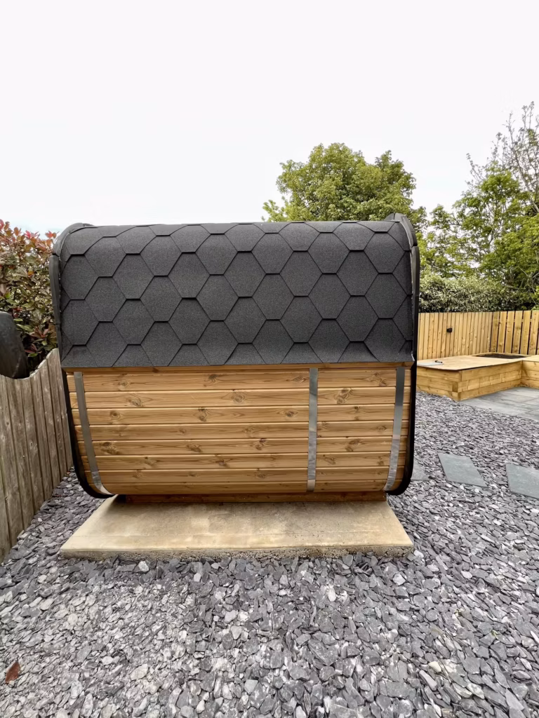 A direct side-profile view of the Yakia 200L outdoor sauna, highlighting the curved rectangular Thermowood timber walls, stainless steel tightening bands, and black bitumen shingle roof cladding.