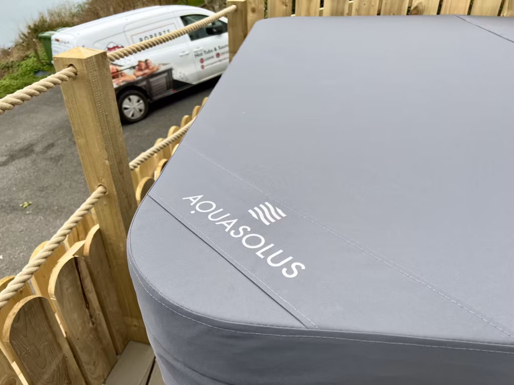 A close-up of a grey insulating cover on an AquaSolus hot tub featuring the white brand logo, with a wooden fence and a local delivery van in the background on the Isle of Man.