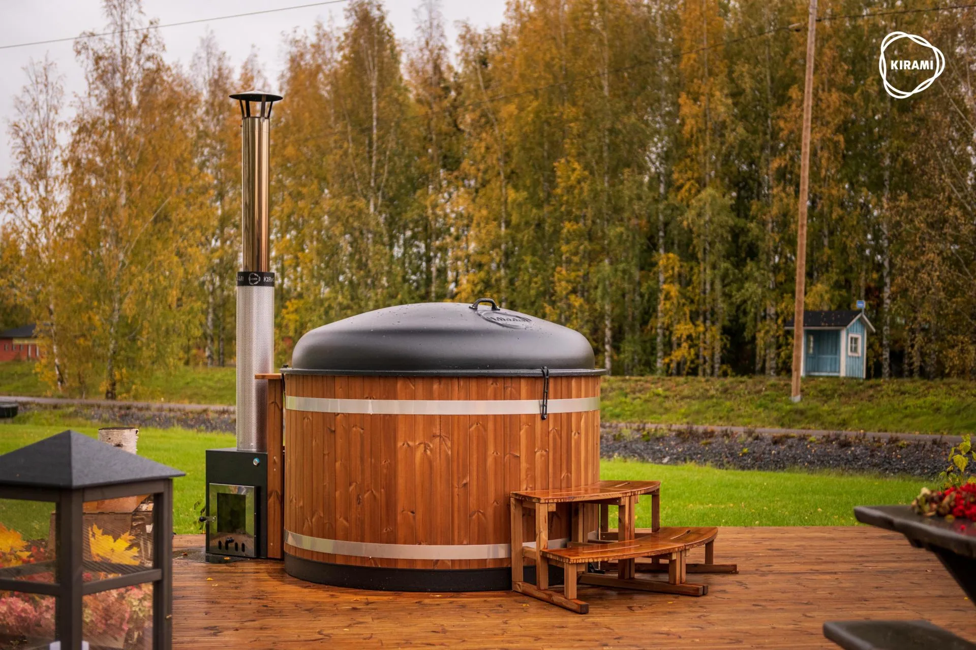 A Kirami Comfort Steady M wood-fired hot tub with a light wood exterior, fitted with a black dome cover and an external heater stove, installed on a spacious wooden deck with autumn trees in the background on the Isle of Man.