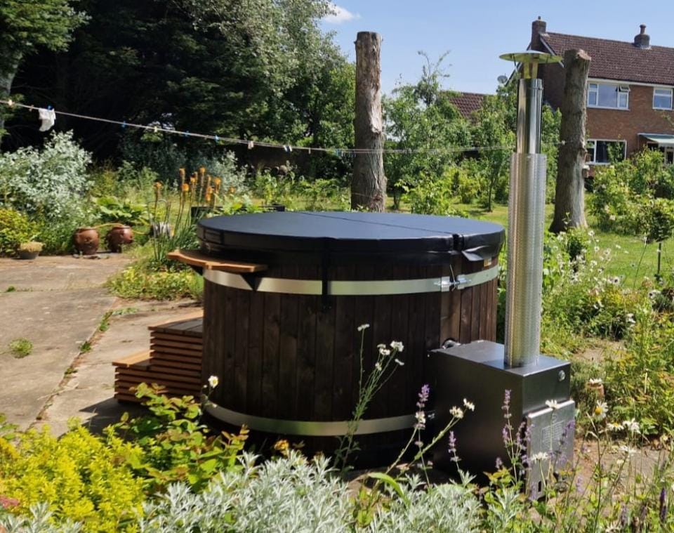A round Kirami Original Hazy M wood-fired hot tub with dark wood paneling and a black cover, installed on a patio surrounded by green foliage and flowers in an Isle of Man garden.
