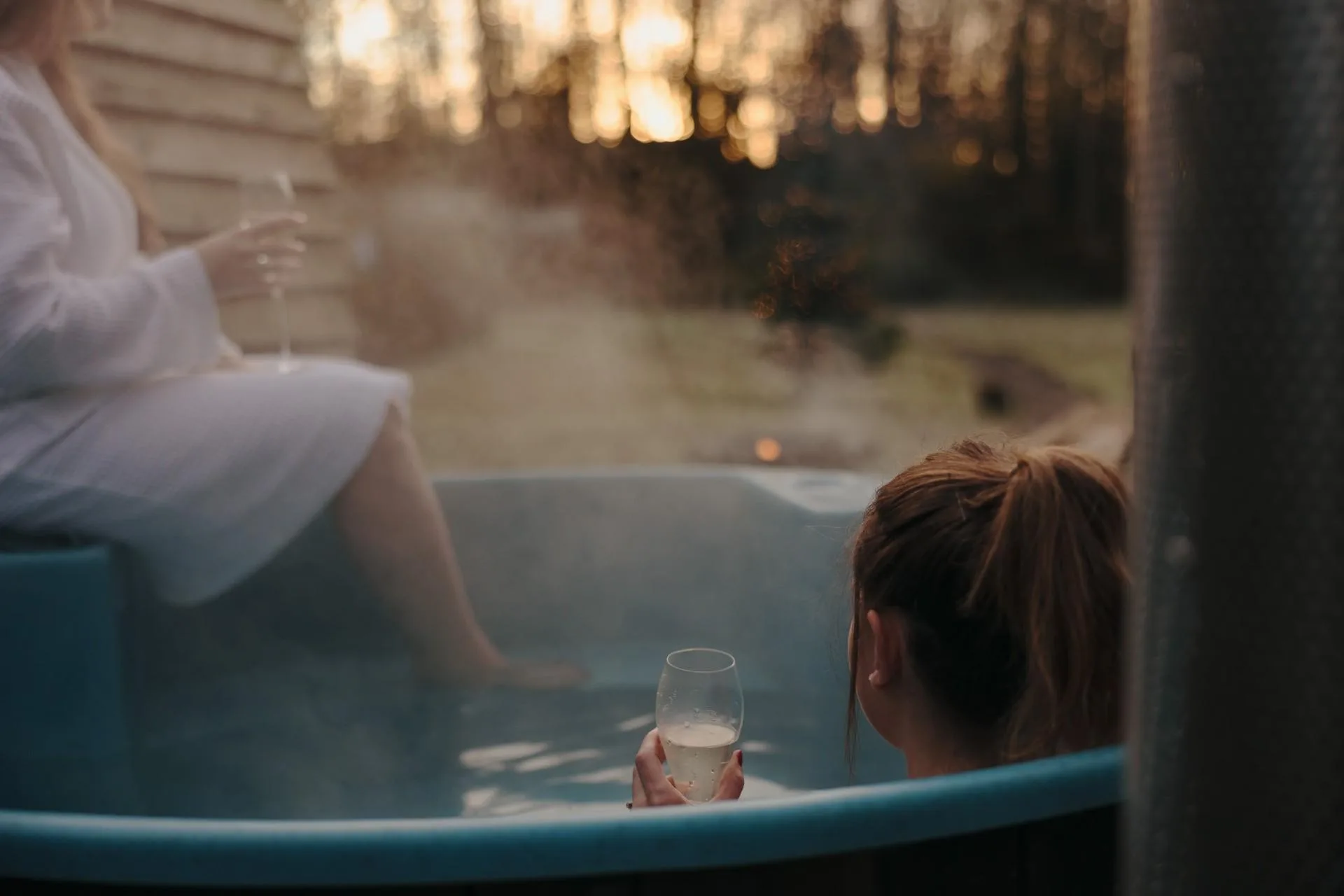 A close-up over-the-shoulder view of a woman holding a glass of sparkling wine while soaking in a steaming Kirami wood-fired hot tub at sunset, with another person sitting on the edge in a white bathrobe on the Isle of Man.