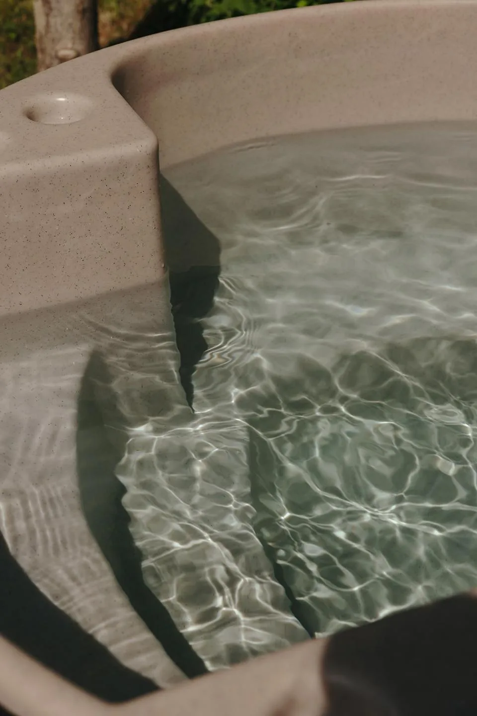 A close-up shot of the clear, rippling water inside a beige molded Kirami wood-fired hot tub, highlighting the integrated safety steps and premium speckled texture liner on the Isle of Man.